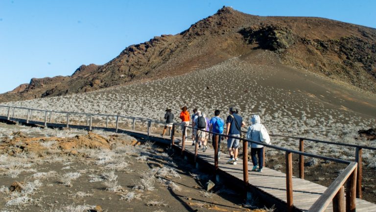 Group of travelers on a Horizon Galapagos cruise walks a boardwalk over an arid zone with a brown peak in the distance.