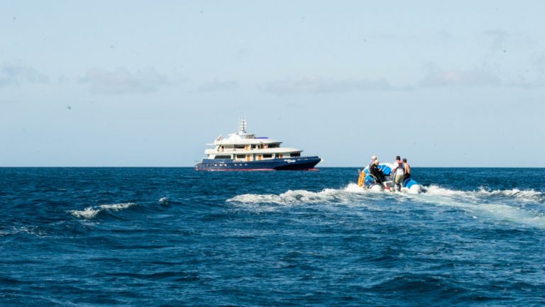 Small ship with red & blue hull & white upper decks waits for travelers approaching on a Zodiac on a Horizon Galapagos cruise.