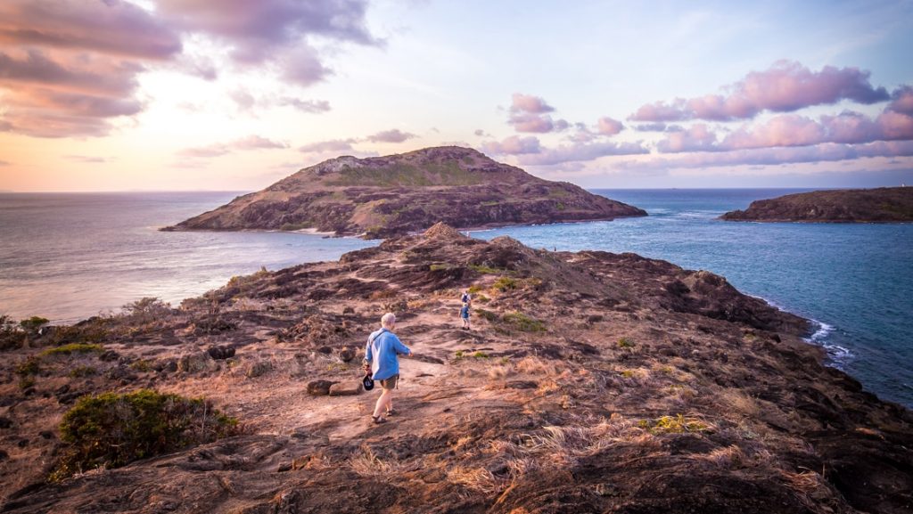 Australia travelers hike along the rocky, sandy top of an island at sunset with purple clouds above & blue sea below.