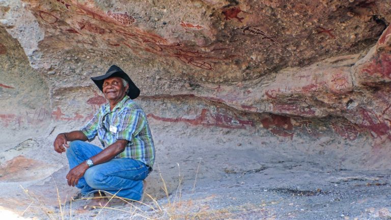 Man in dark cowboy hat, checkered shirt & jeans kneels down & smiles beneath indigenous rock art, seen in Arnhem Land.