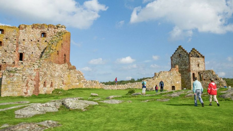 2 travelers walk toward old brick castles atop bright green grass under a blue sky on the Circumnavigating the Baltic Sea Cruise.