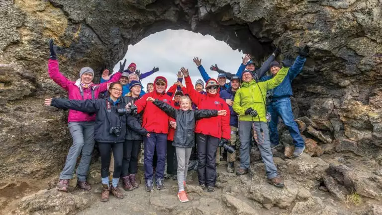 Group of Iceland Circumnavigation cruise guests stands with arms outstretched for a photo opp under a gray lava arch.