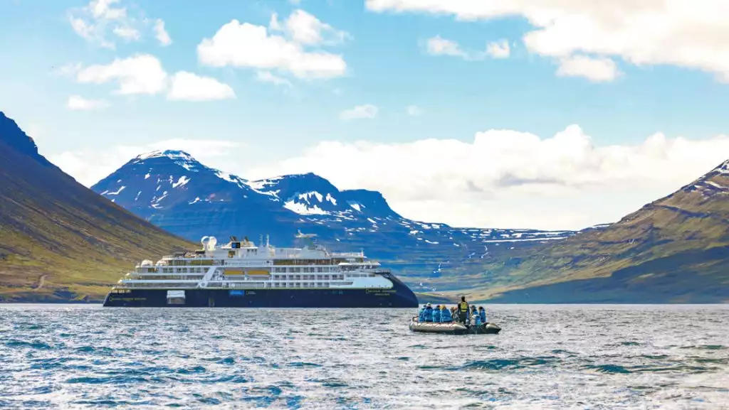 Nat Geo small ship in blue & white sits offshore from Icelandic mountains of green, brown & snowy peaks, as Zodiac boat nears.