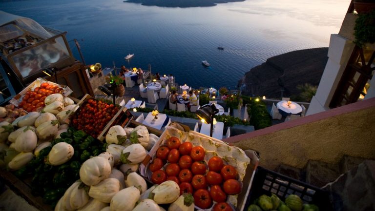 Vegetables laid out in boxes look down on travelers dining on a terrace overlooking calm seas at sunset in Greece.