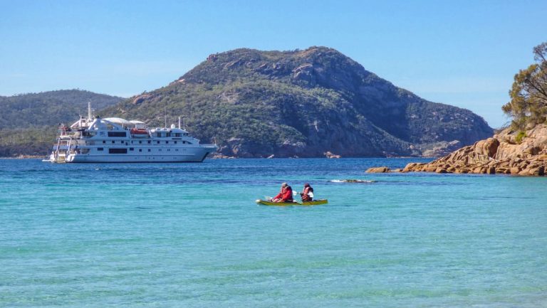 1 tandem kayak with Tasmania travelers moves through calm turquoise water on a sunny day with a white small ship in the background.