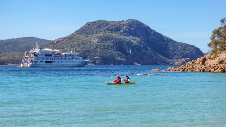 1 tandem kayak with Tasmania travelers moves through calm turquoise water on a sunny day with a white small ship in the background.