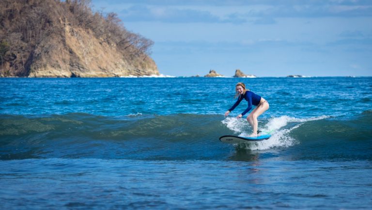 Girl surfs a small wave in bright blue ocean with a tan cliff behind on a sunny day during the Costa Rica Family Adventure.