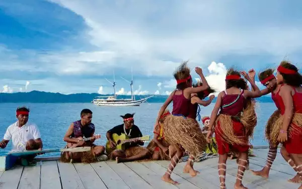 Small cruise ship seen behind colorfully dressed dancers and cruise guides seated playing instruments on a dock in Raja Ampat Indonesia