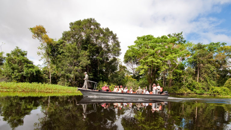 group of small ship passengers cruises down the amazon in skiff on a sunny day with trees behind them with a guide standing at the front
