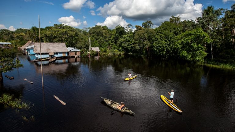 Aerial view of 2 stand-up paddlers & a solo kayaker beside rustic huts over the water & dense Amazon rainforest beside.