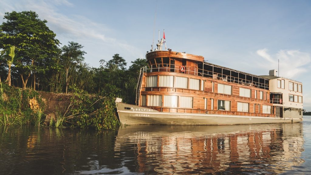 Delfin II Amazon river boat sits in glassy water beside the riverbank, with 3 wood-wrapped decks & a single sage green hull.