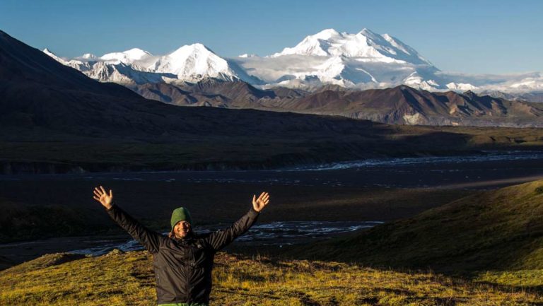 Man smiling with his arms up in the air with a river and mountain range behind him in alaska's denali national park