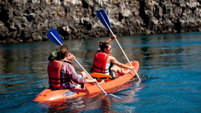 Tandem kayaking pair paddles an orange boat beside rocky cliffs in calm water on an Evolution Galapagos cruise.