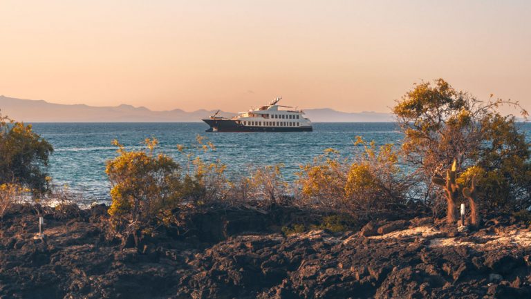 Evolve Galapagos yacht with dark blue hull & 2 white upper decks sits in calm water by rocky small island at sunset.