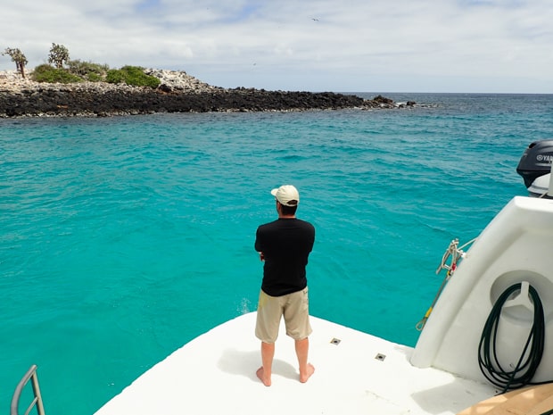 A man standing on the stern of a Galapagos small ship looking at the turquoise water and a nearby island