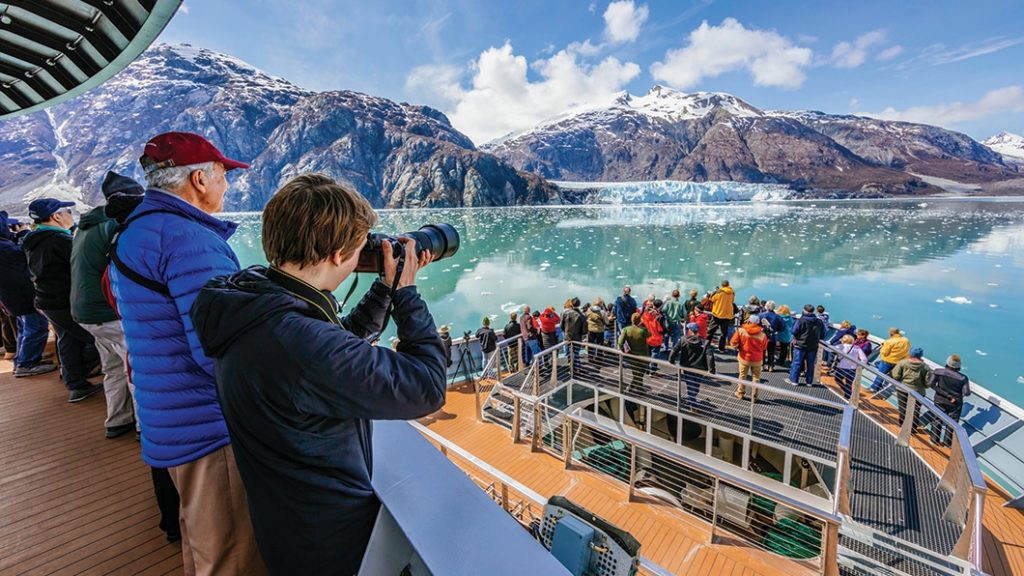 Cruise guests gather at the ships bow to take photos of a large glacier that extends to the water from an Alaska mountain range.