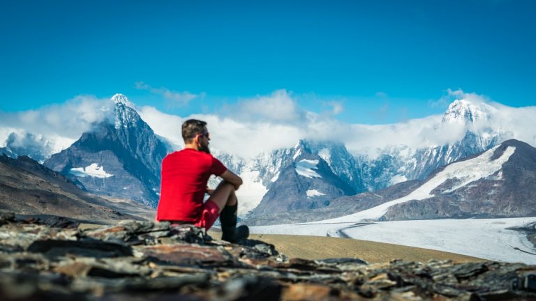 Man in bright red shirt sits atop a mountainside, looking out over snowcapped peaks on a sunny day in Antarctica.