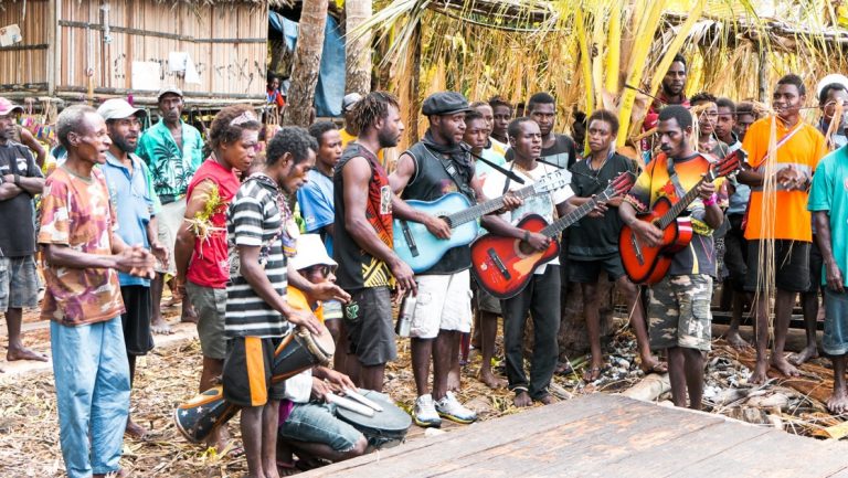 Local men stand and play drums & guitars while singing among thatch buildings, seen on a Papua New Guinea cruise.
