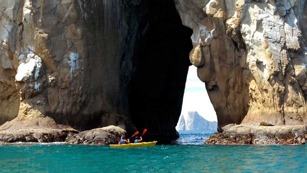 2 travelers paddle a yellow double kayak through the arch of a giant rock structure in the Galapagos, in the distance another iconic rock landmark, Kicker Rock.