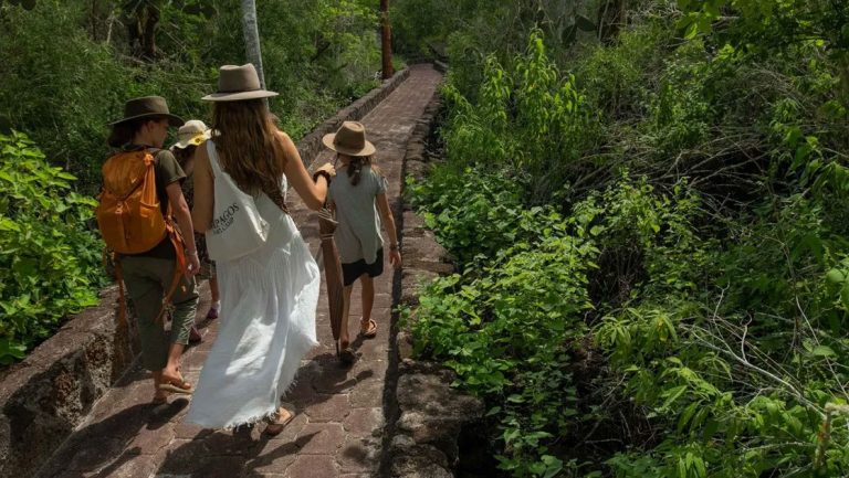 Woman in felt hat & white sundress walks with 3 kids on a stone pathway among bright green jungle during a Galapagos Safari.