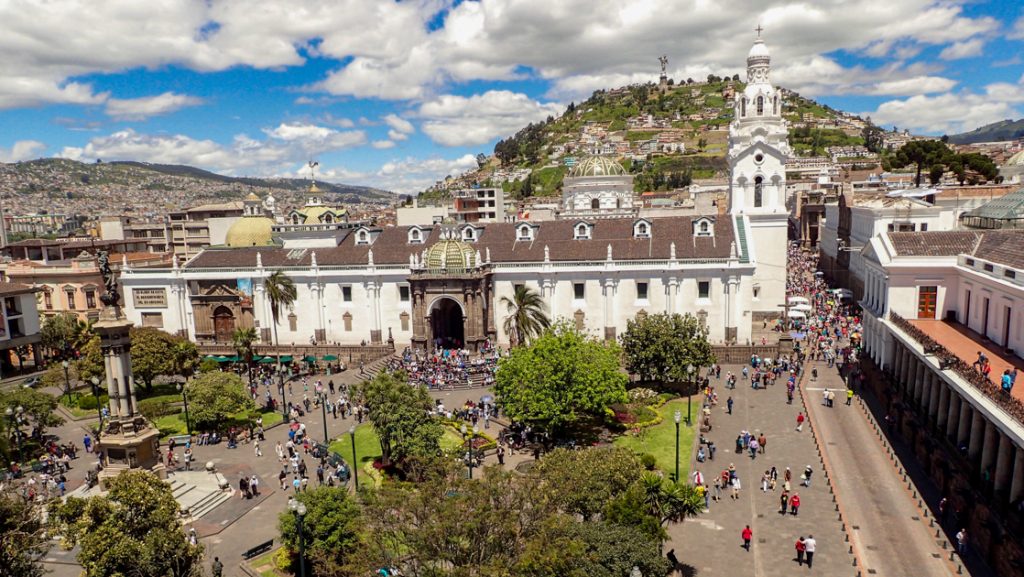 Aerial view onto city square with historic governmental buildings, bright green grass & trees & a lot of pedestrians in Quito.