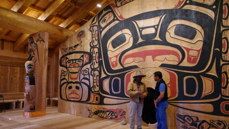 two men in a wooden tribal house with carvings and totem paintings in alaska