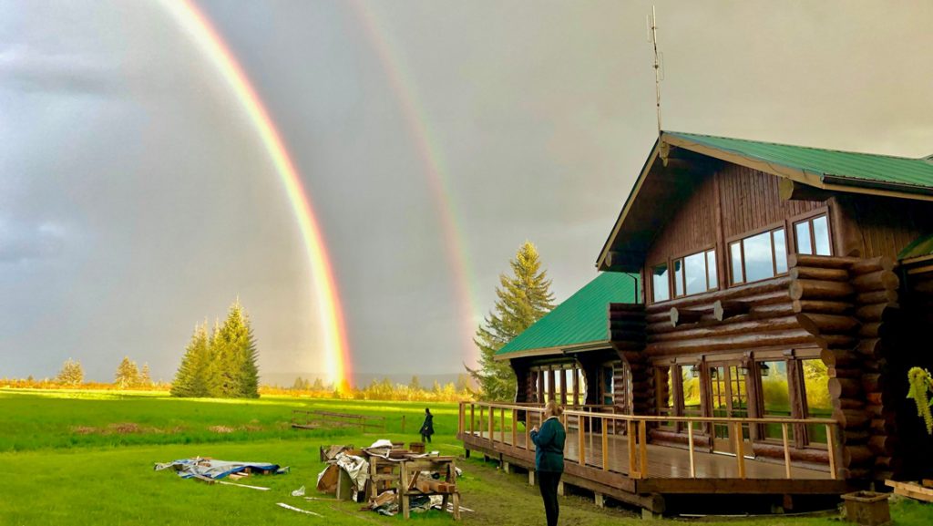 Woman stands outside log cabin lodge in Alaska & views a double rainbow over a grassy field in the morning light.