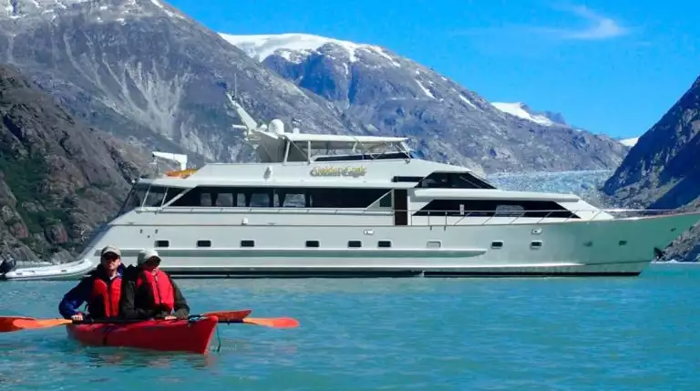 Two guests from the Golden Eagle yacht paddling in a double kayak in front of their small ship on a sunny day in Alaska.
