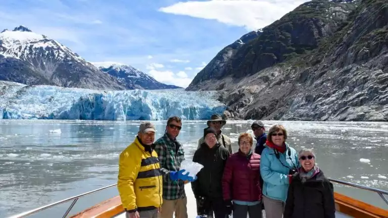 Group of adventure travelers on the bow of the Golden Eagle yacht with a glacier behind them on a sunny day in alaska.