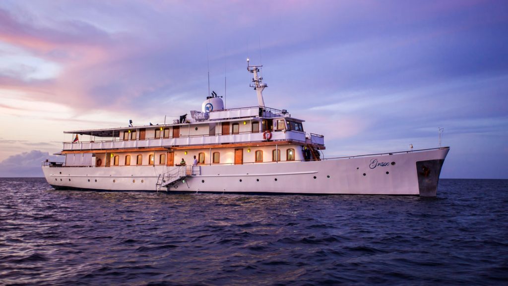 Grace Galapagos cruise ship with white decks & wooden accents cruising in calm waters under purple clouds at sunset.