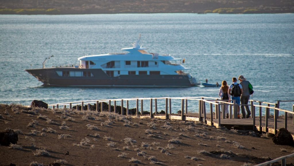The Infinity Galapagos yacht floats off shore as cruise guests walk down a wooden ramp to get back on board after a shore excursion.