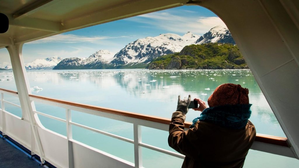 From the deck of a ship a guest holds a camera up to take a photo of the Alaska landscape while aboard Inside Passage Sojourn Alaska cruise.