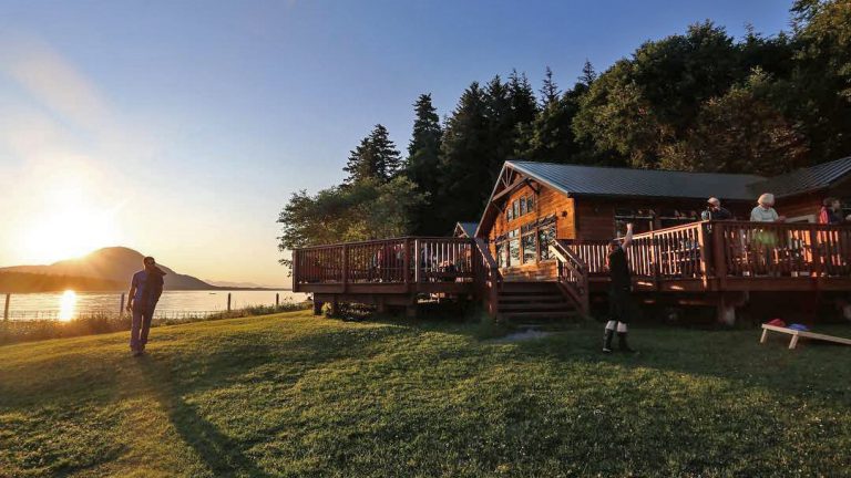 The sun is setting over the mountain range that surrounds the Orca Point lodge in Alaska as guests enjoy playing games on the lawn and patio.