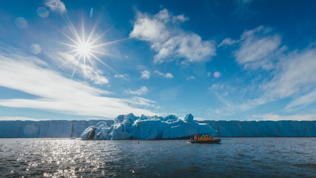 Zodiac boat with Arctic travelers in yellow jackets cruises past 1 long ice shelf & smaller iceberg in front on a sunny day.
