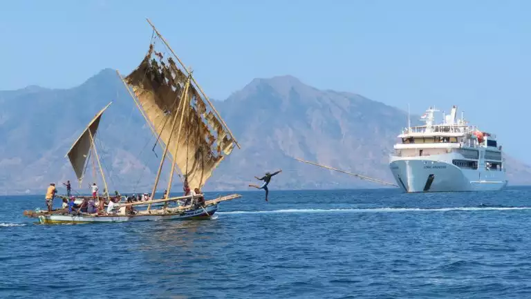 Small white ship sits nearby tiny fishing boat with weathered sails as man jumps off bow throwing a spear in Indonesia.