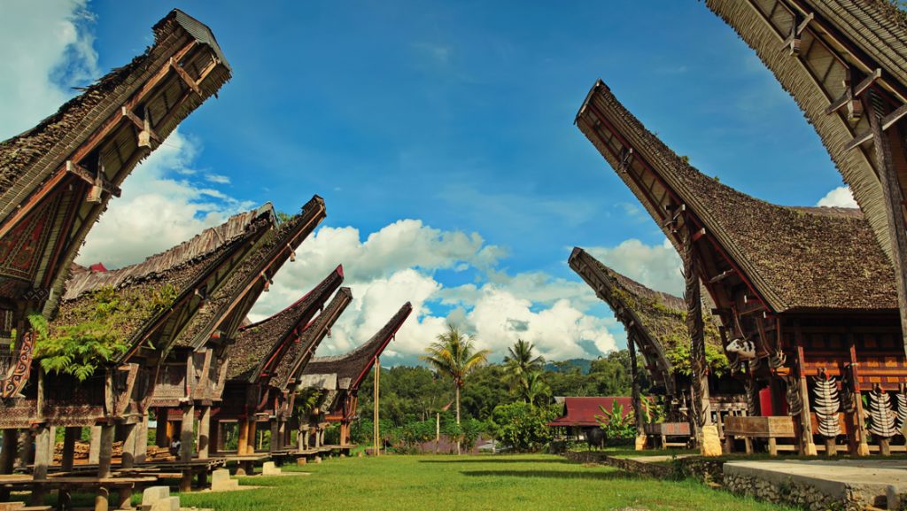 Row of thatched luxury bungalows with tall, pointed roofs facing each other across the grass on a sunny day in Indonesia.