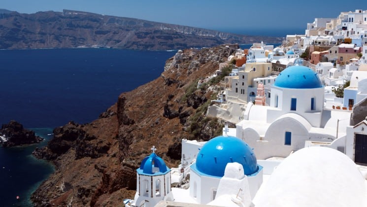 Aerial view of the white-stone buildings with blue-domed tops sitting on a hillside overlooking deep blue water in Santorini, Greece.