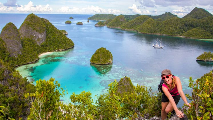 a solo female traveler stands smiling at the top of a peak with a beautiful indonesia cove below her and a small ship anchored in the middle of the water