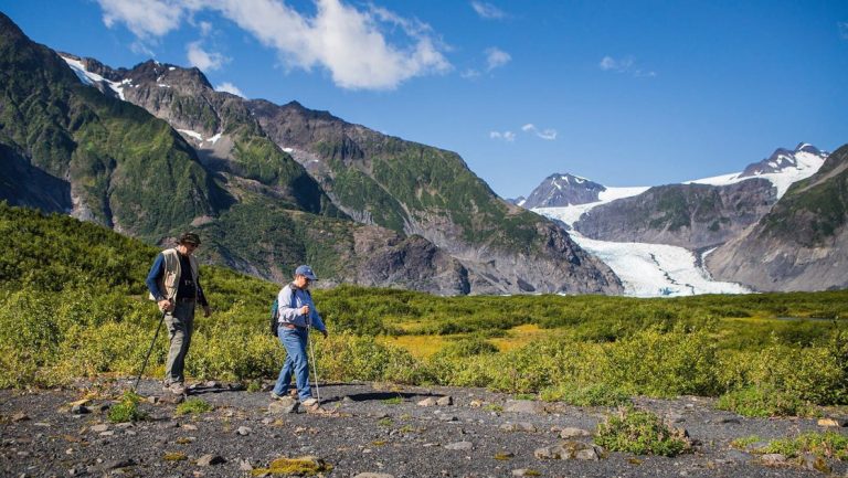 2 men hike over flat terrain with small rocks with a glacier, rocky mountain peaks & green shrubs behind in Kenai Fjords NP.