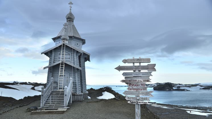 an old white church stands alone on an antarctica shoreline with a signpost in front of it and dark clouds above