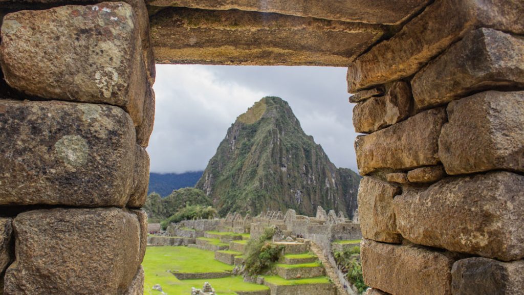 View through opening in brown stone wall at Machu Picchu citadel, looking onto bright green grassy levels & tall mountain behind.