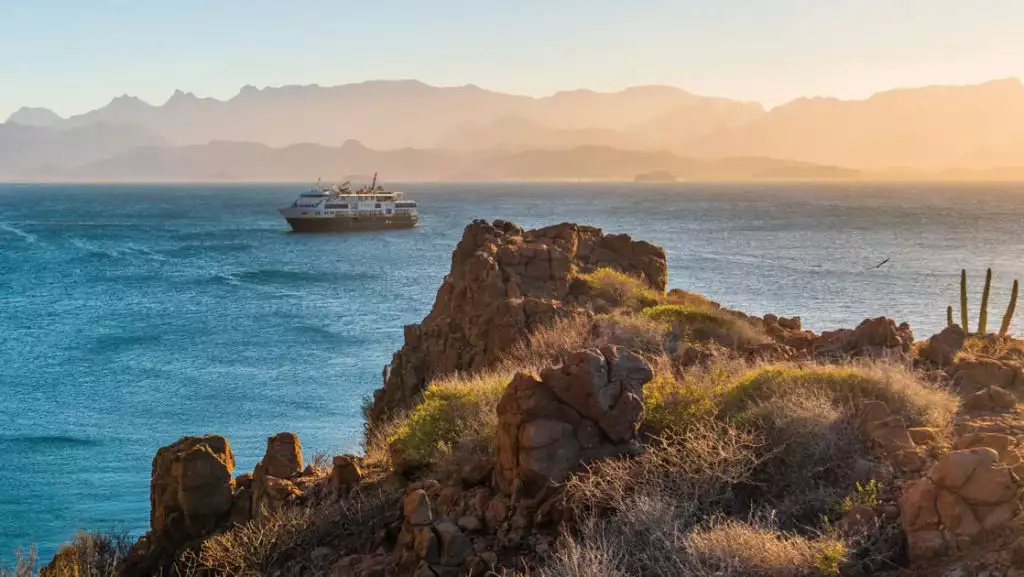 National Geographic small ship sits offshore of rocky Baja coastline with green & gold shrubs, at sunset.