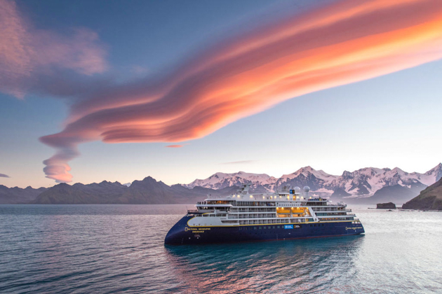 Small polar ship with blue hull & white upper decks sits under pink & purple clouds on the Nat Geo White Continent cruise.