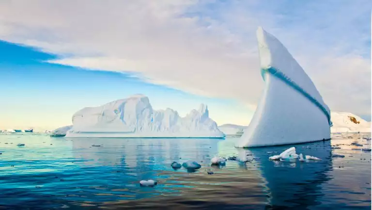 Large, skinny, rounded iceberg rises like a wall out of calm turquoise water, seen on the Nat Geo White Continent cruise.