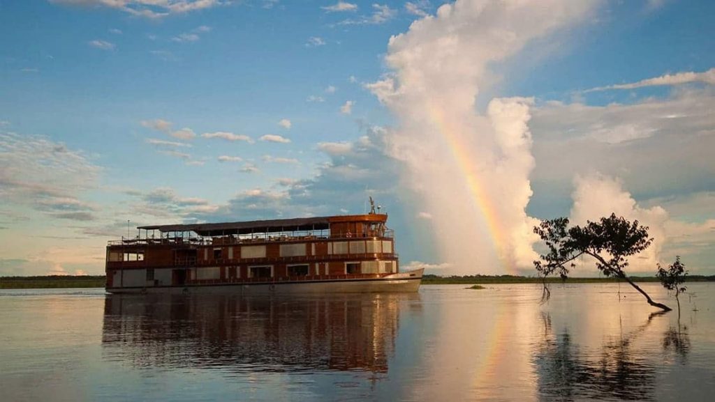Delfin II amazon river boat cruising on calm water with a bright cloud and rainbow behind it.