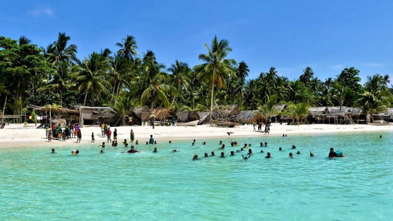 Locals swim in turquoise water beside a white-sand beach with thatch huts & palm trees, seen on New Guinea cruises.