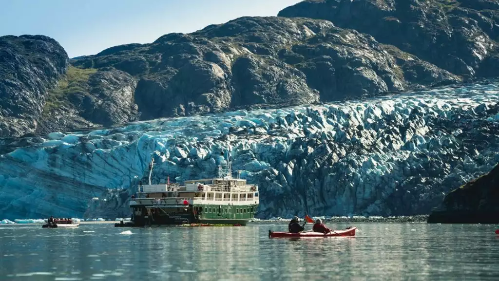 The white and green small ship the Wilderness Adventurer floats in front of a massive glacier in Alaska as kayakers paddle around it.