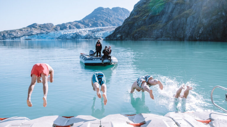 Guests leaping off the kayak towards zodiac platform into glacier bay for a polar plunge on a bright sunny day