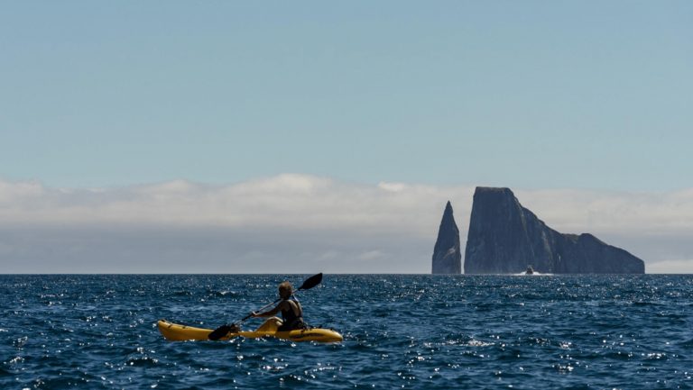 Kayaker in Galapagos paddles a yellow boat in choppy water on a sunny day with a large rock island in the distance.