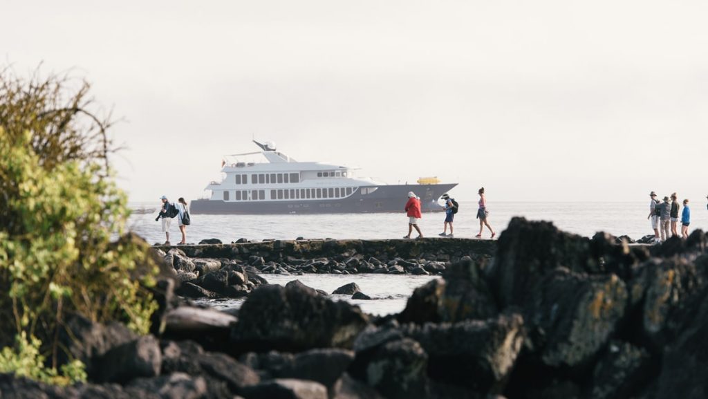 The luxury Origin and Theory Galapagos yacht floats beyond the shore line as cruise guests walk along the black lava rock on shore.